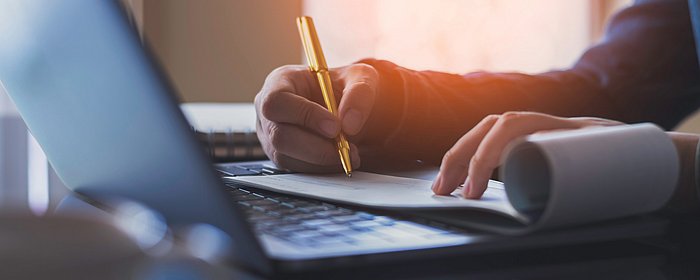 Eine Person schreibt im Notizbuch, am Laptop, links unscharf eine Kaffeetasse 