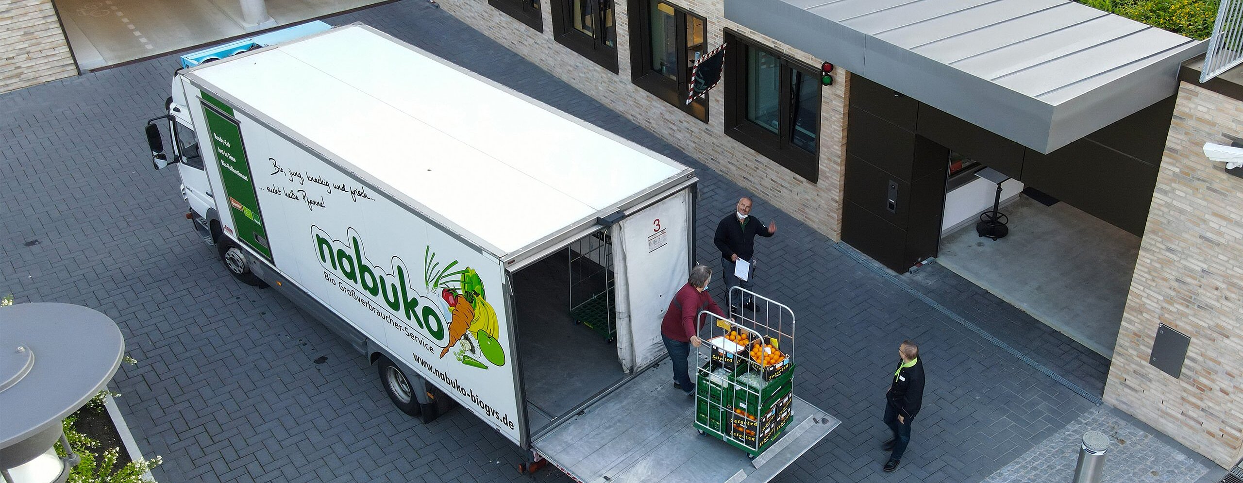 Ein beige, graues Firmengelände. Im Innenhof ein weißer LKW der Firma nabuko. Diese laden Obst und Gemüse aus 