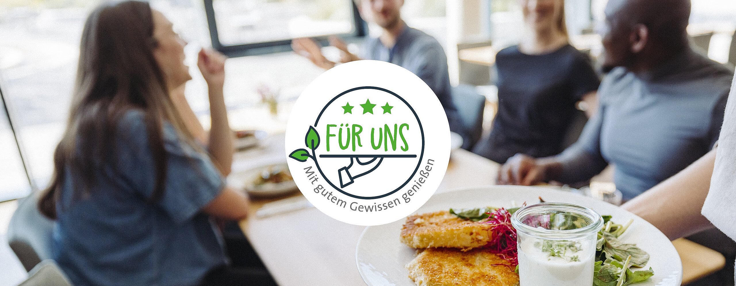 Im Hintergrund eine Gruppe am Mittagstisch, mittig das "FÜR UNS" Siegel, rechts darunter- ein weißer Teller mit Schnitzel und Salat