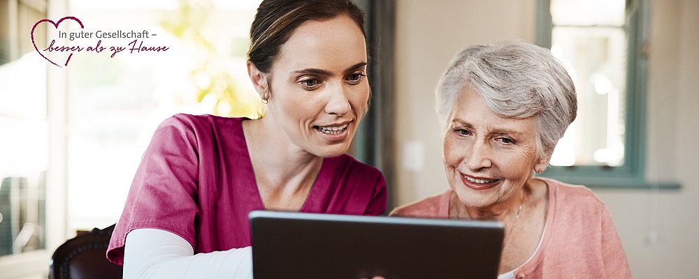 Krankenpflegerin in bordeaux rotem Kittel zeigt älterer Damen etwas auf dem Ipad. 