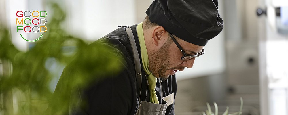 "Ein konzentrierter Koch in schwarzer Kochmütze und Schürze bereitet in einer modernen Küche eine Speise zu unter dem GOOD MOOD FOOD Logo
