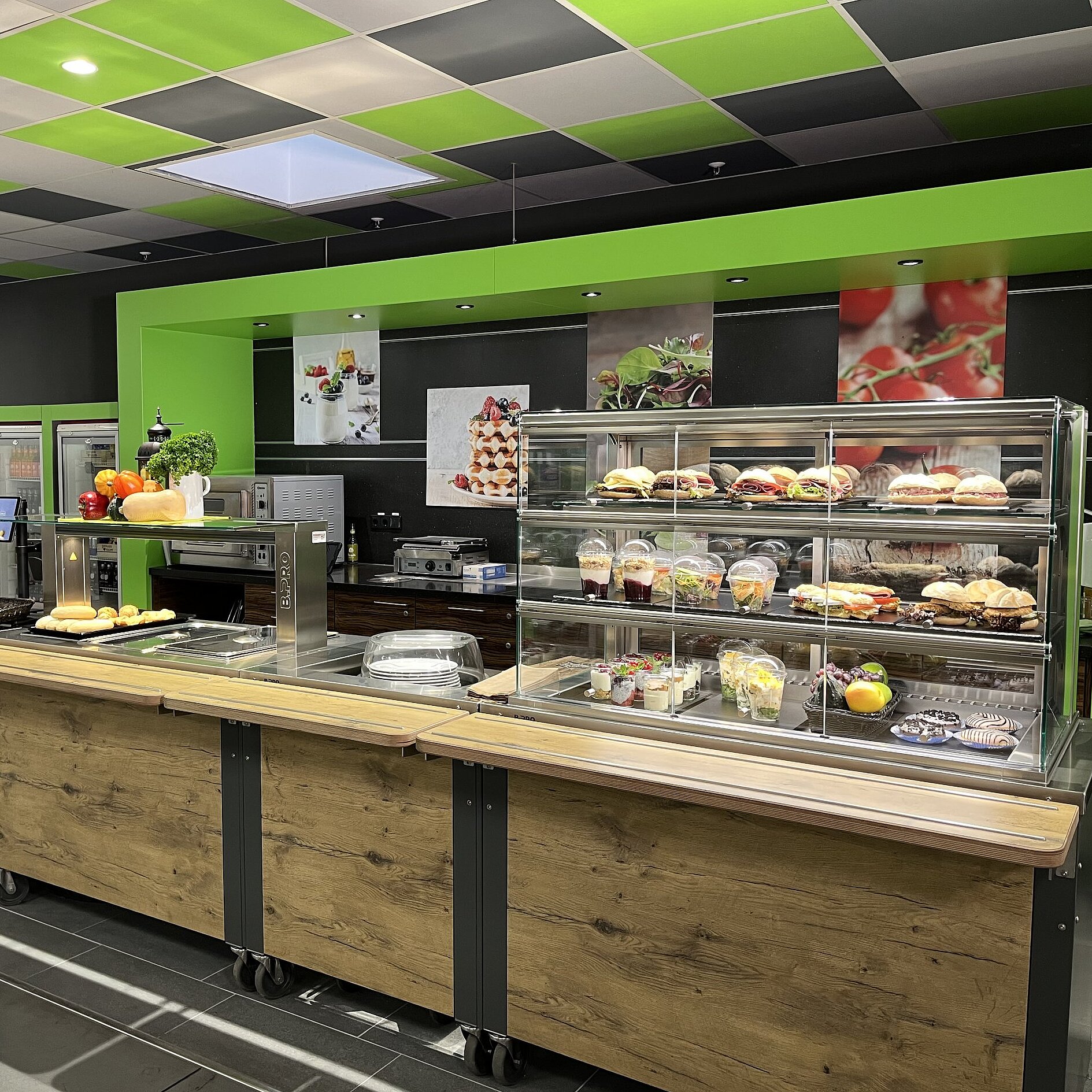 Eine holzfarbige Auslage gefüllt mit Obst und Backwaren. Im Hintergrund eine hellgrüne Umrandung, darin schwarze Fliesen.  