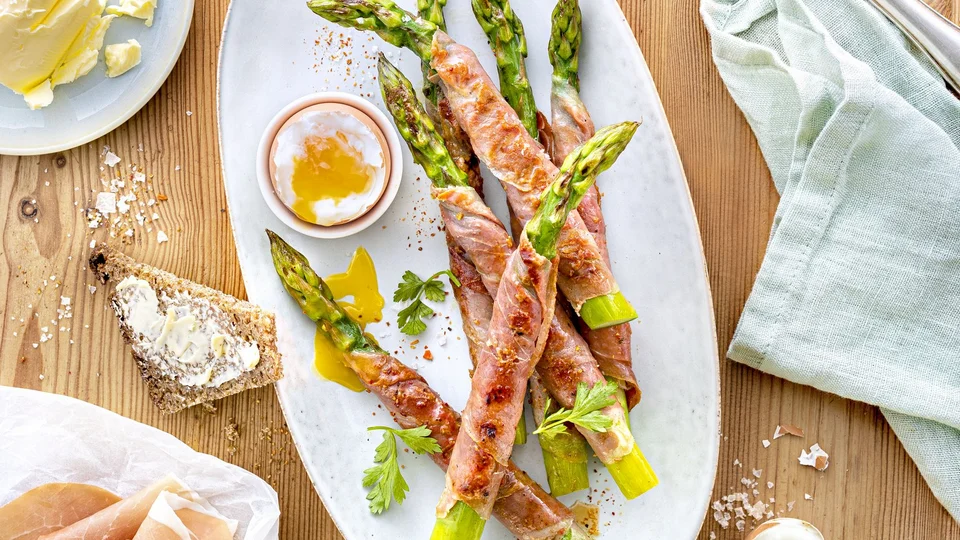 Mittig ein oval weißer Teller, darauf grüner Spargel in Schinken eingewickelt. Oben links Vollkornbrot darauf gelegt ein Messer. Linksseitig ein Stück Butter und Schinken. Alles drapiert auf einem hellbrauen Tisch